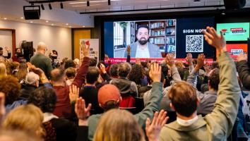 Audience members raise their hands at the “Justice for Palestine” European Citizens’ Initiative (ECI) event, watching a large screen showing a man speaking remotely, with campaign messages and a QR code displayed beside him.