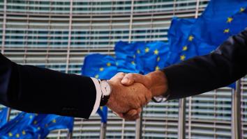 Two people in business suits shaking hands in front of several European Union flags in front of the EU Commission building