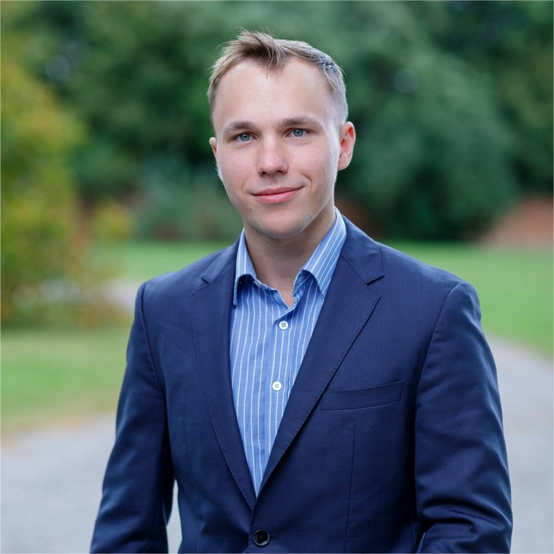 “Portrait of a young adult wearing a navy blue blazer and a light blue striped shirt, standing outdoors on a pathway with green trees blurred in the background.”