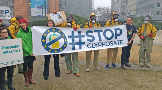 A group of people stands outdoors during a public demonstration, in front of the European Commission building in Brussels, holding a large banner that reads “#STOP GLYPHOSATE,” featuring a crossed-out pesticide sprayer symbol inside a circle of European Union stars. Some individuals wear protective suits and gas masks, while others are dressed casually. One person holds a sign reading “Their profits – our cancers.”