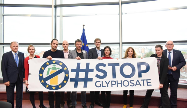 Organisers of the “Ban Glyphosate” European Citizens’ Initiative hold a large banner reading “#STOP GLYPHOSATE,” featuring a crossed-out pesticide sprayer symbol within a circle of European Union stars. They stand alongside an EU Commissioner and other institutional representatives in a formal setting at the European Commission.