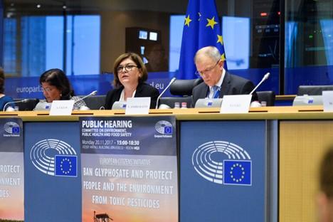 Three speakers sit behind a long desk in a conference room at the European Parliament, facing an audience. They speak into microphones while consulting documents or screens. Below them, a large banner reads “Public Hearing: Ban Glyphosate and protect people and the environment from toxic pesticides.” The setting is a formal European Union event, with EU logos and flags visible in the background.