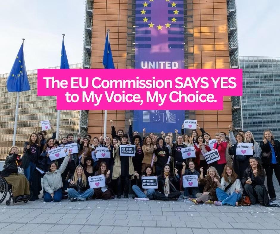 A large group of activists of My Voice My Choice celebrating in front of the European Commission Berlaymont building in Brussels, with a prominent text overlay announcing the major campaign victory.