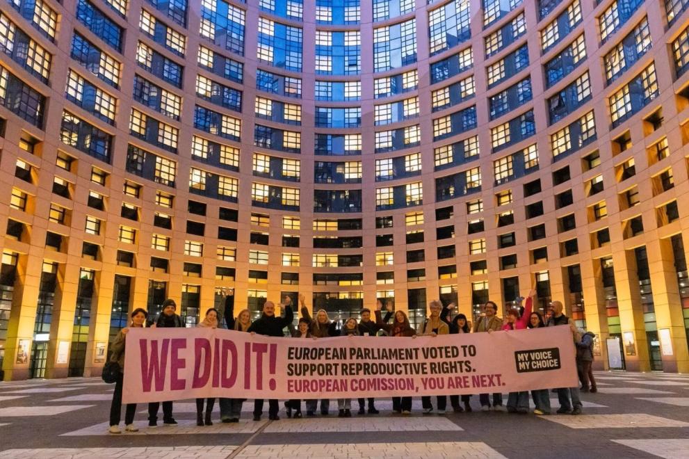 The group of activists of My Voice My Choice standing in the main round square of the European Parliament in Strasbourg, holding a large horizontal banner to celebrate a successful campaign milestone.