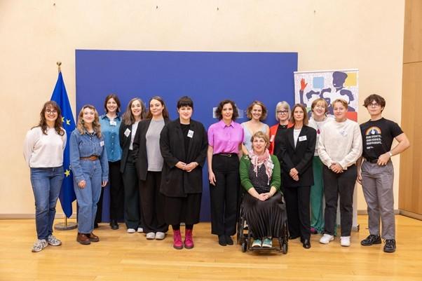 A group portrait of fifteen activists and organizers standing in front of a blue backdrop and an EU flag, representing a diverse team of campaigners.